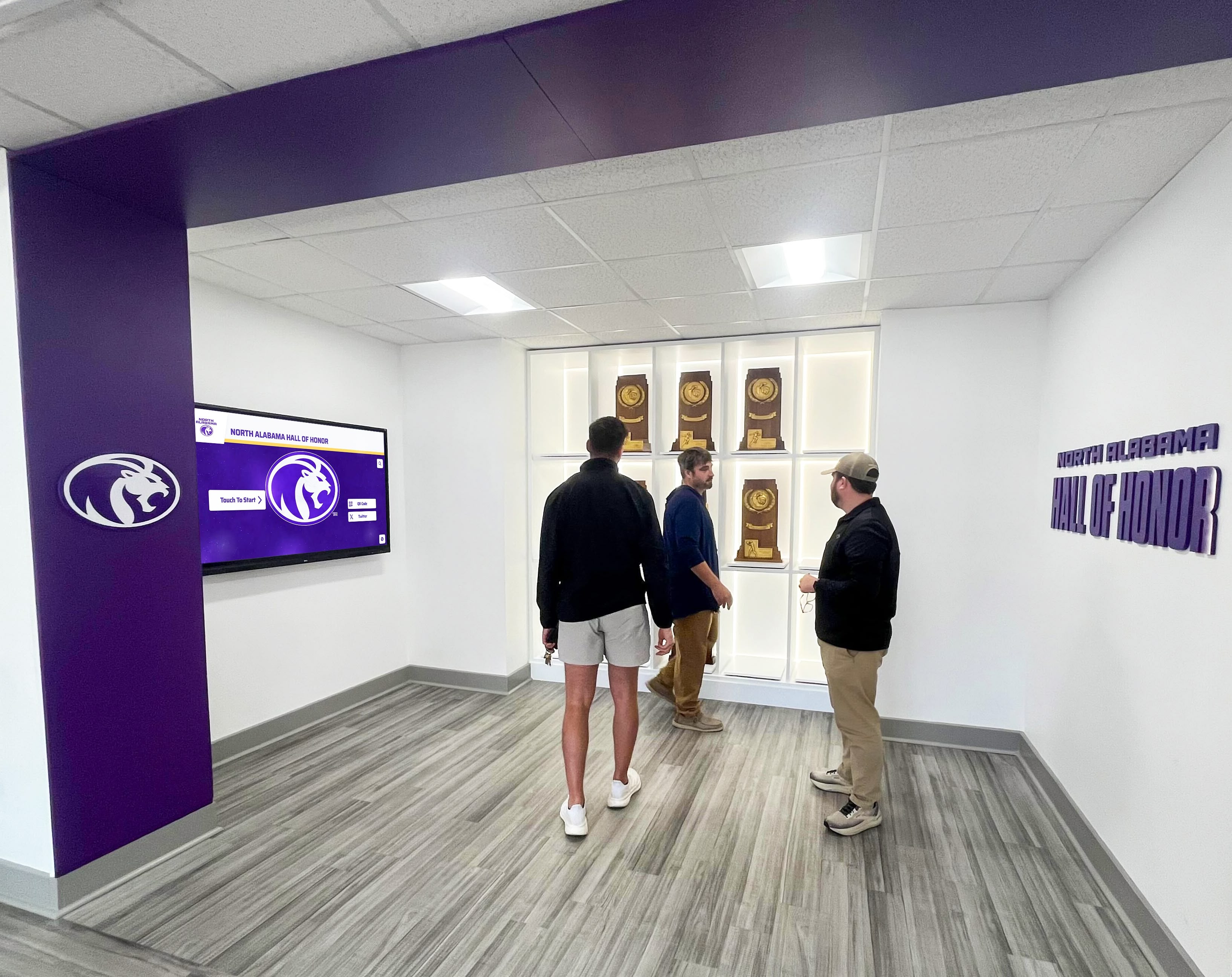 Three people viewing hall of honor trophy display Multiple people viewing digital athletic recognition